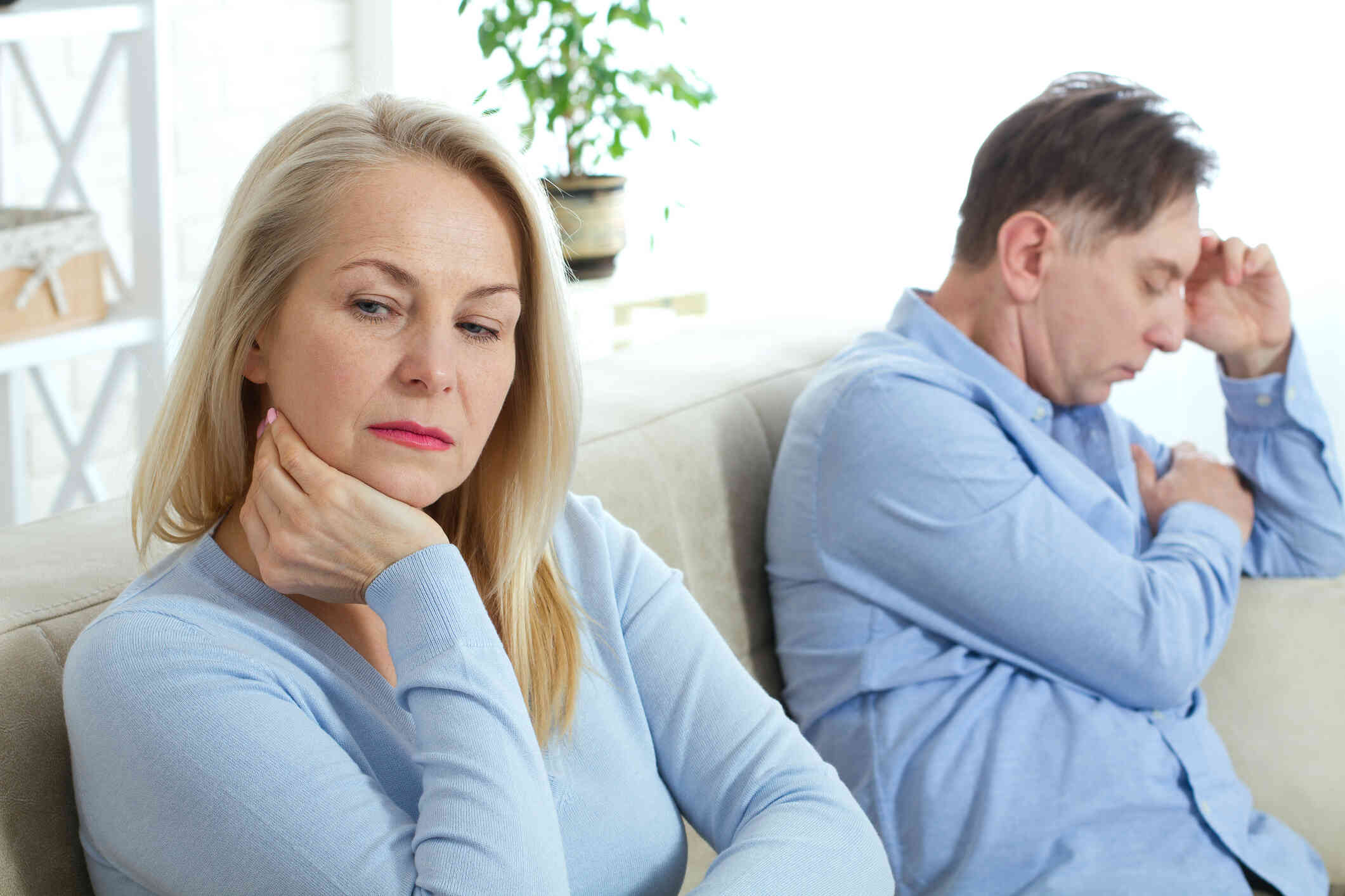 A middle aged couple sit sadly on opposite end of the couch while looking away from each other.